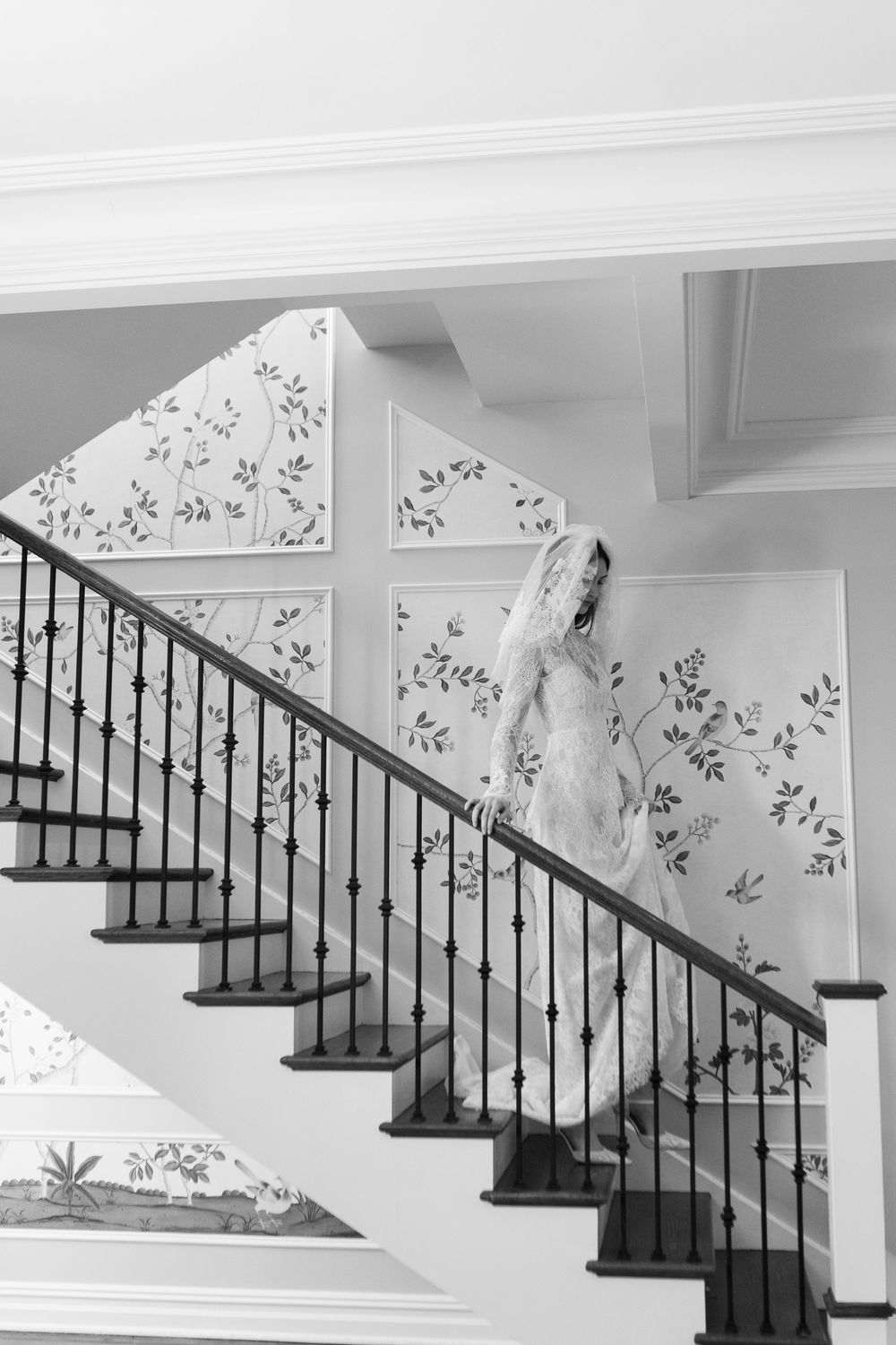 Bride in wedding gown and veil descends elegant staircase with wrought iron railing and floral wallpaper in black and white.