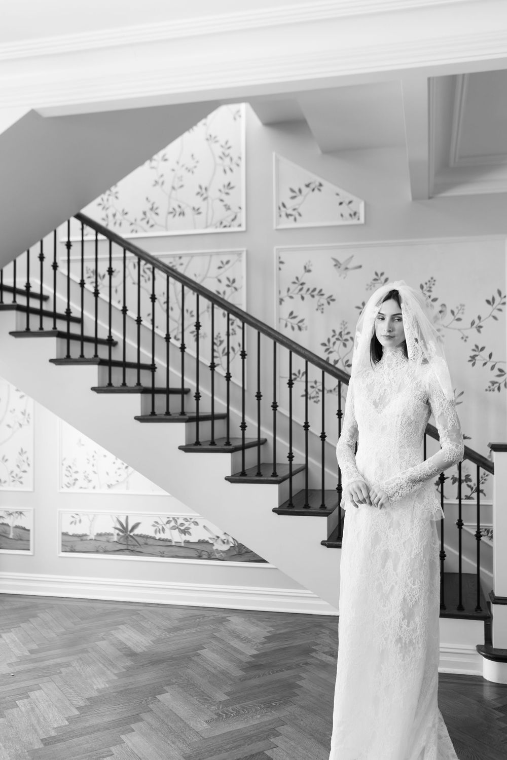 Bride in lace gown and veil posing by a grand staircase with floral wallpaper.