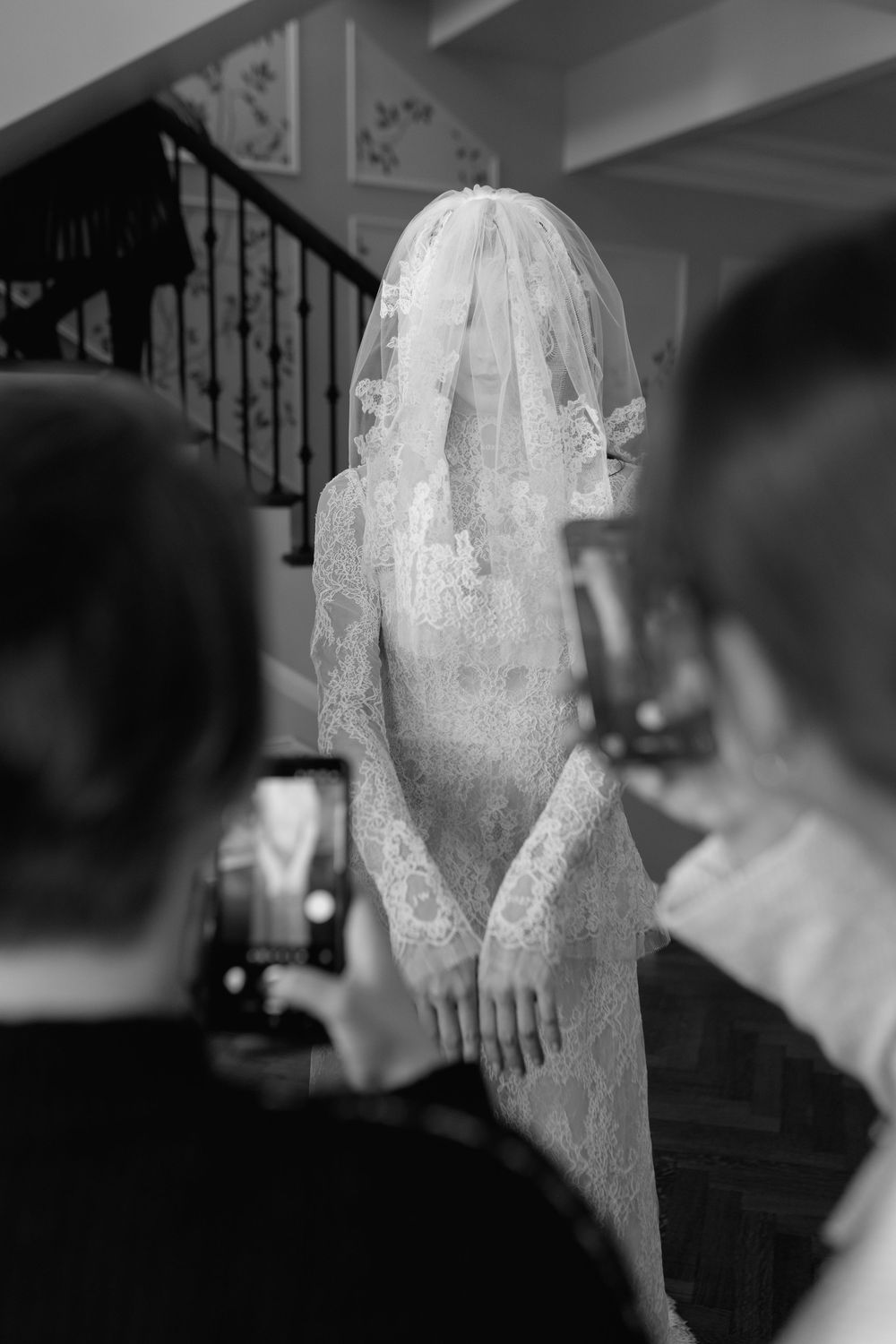 Black and white photo of a bride in lace gown and veil, photographed by guests on smartphones indoors.