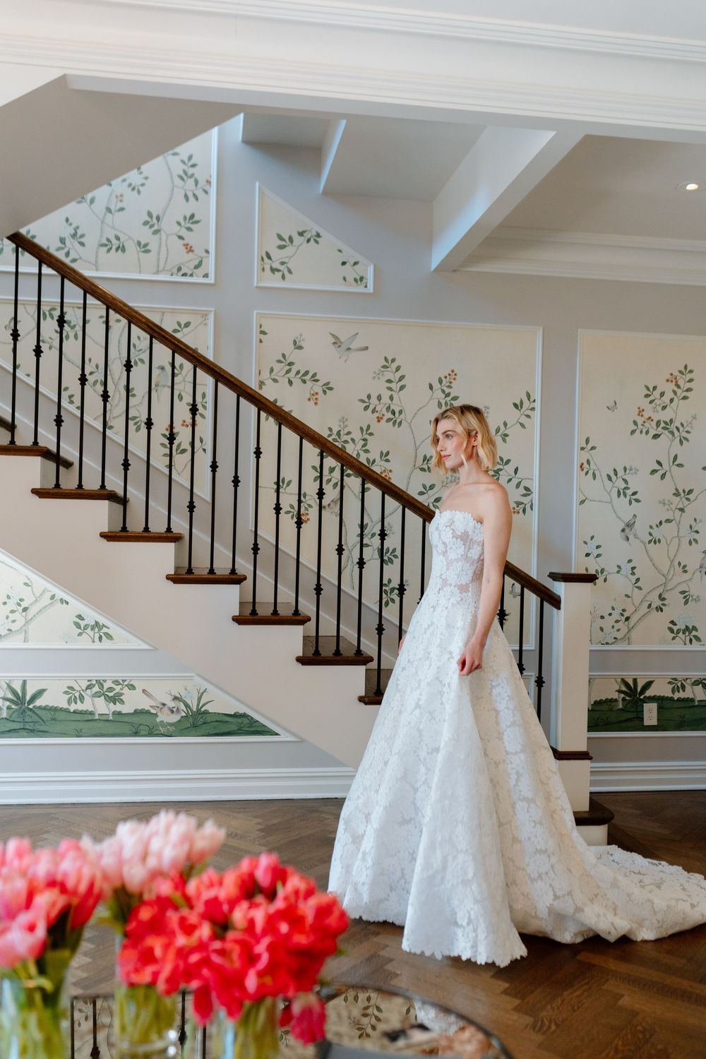 Bride in a stunning lace A-line gown poses by a grand staircase with floral wallpaper.