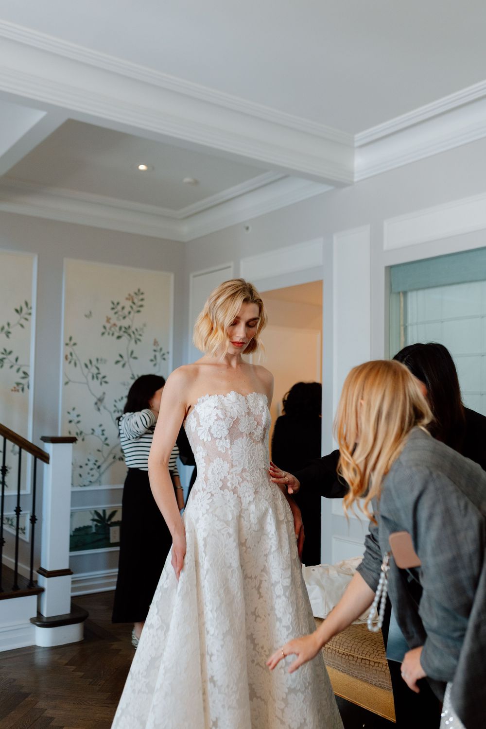 Bride in strapless lace A-line wedding gown being fitted by assistants in an elegant white interior.