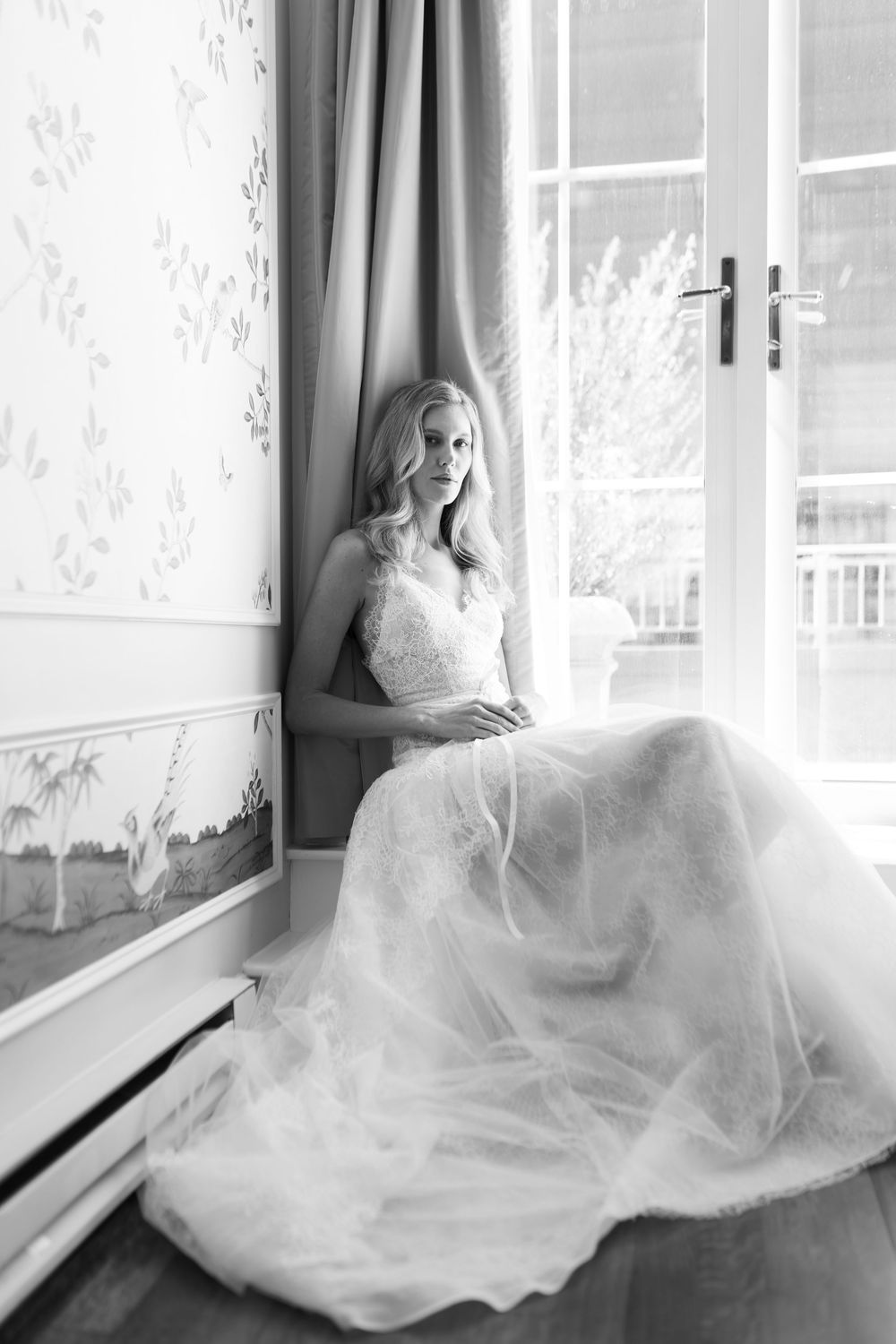 Bride in elegant lace wedding gown sitting by a sunlit window in a classic interior.