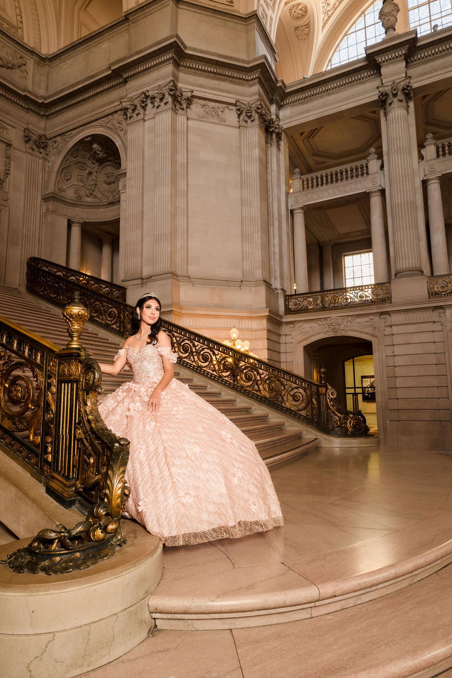 Young woman in a pink ball gown poses on grand staircase in an elegant ornate building.