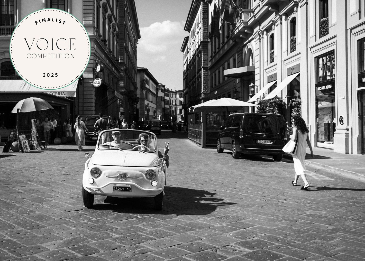 Black and white photo of a vintage Fiat 500 convertible driving through a sunny Italian city street with pedestrians.