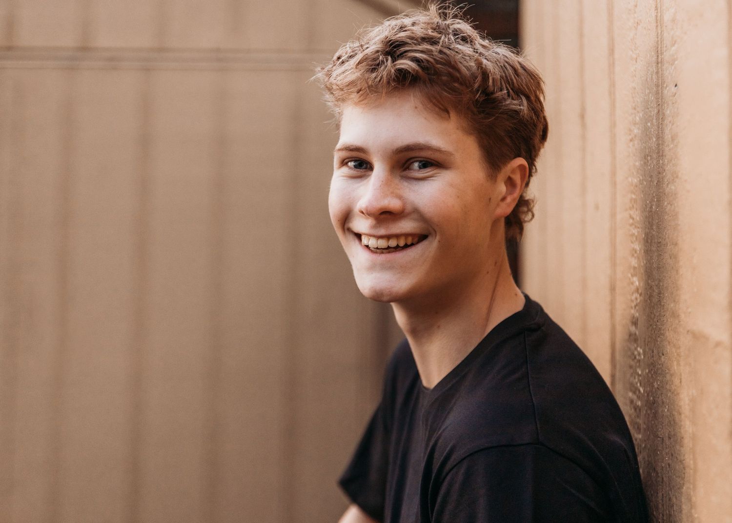 Smiling young man with short hair wearing a black t-shirt, leaning against a wall outdoors.