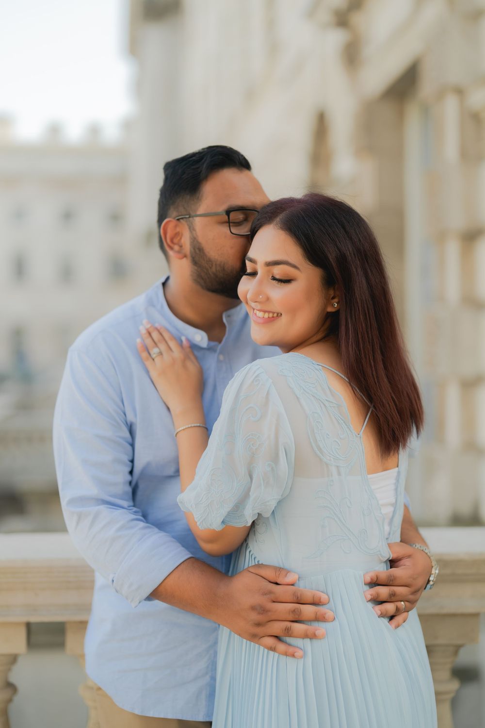 Couple sharing a romantic embrace outdoors, man kissing woman's cheek while she smiles, both in light blue outfits.