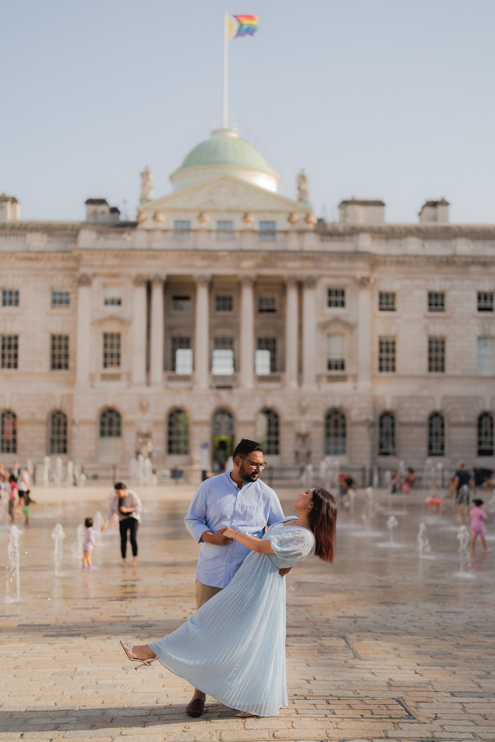 Couple dancing in Somerset House courtyard, London, with rainbow flag and neoclassical architecture backdrop.