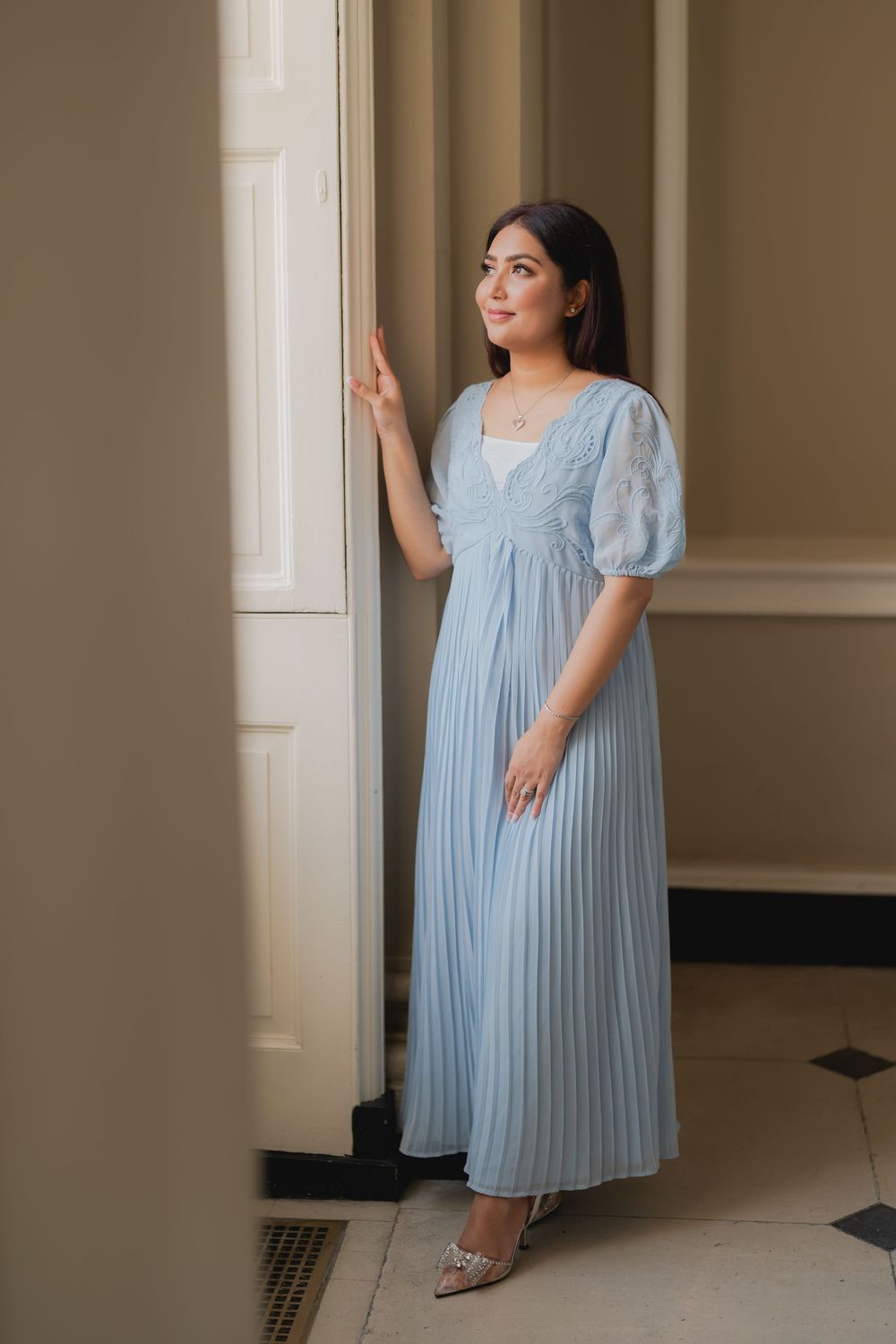 Woman in a light blue pleated maxi dress with lace puff sleeves posing elegantly by a door in a classic interior.