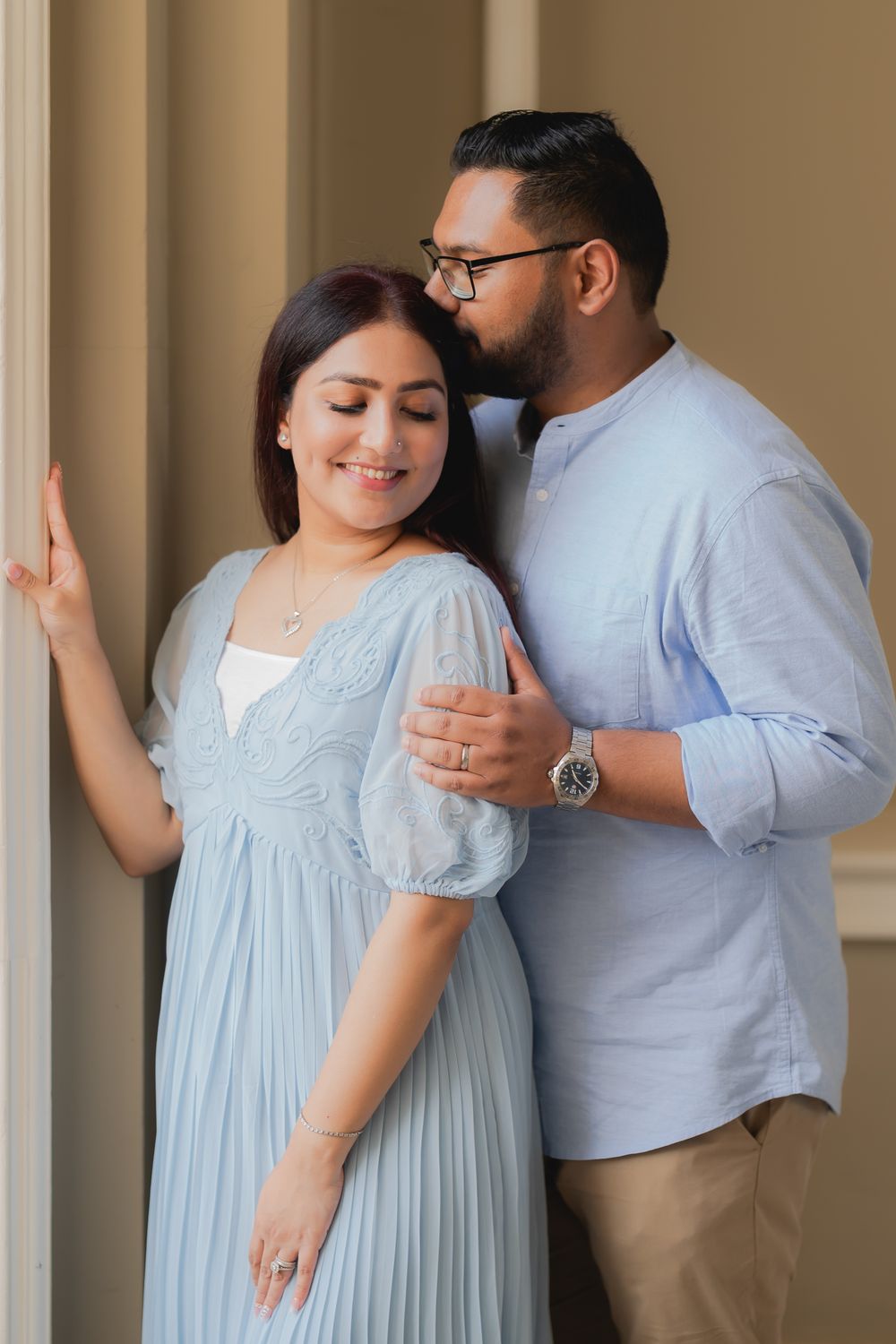 A couple in light blue outfits share a tender moment, the man embracing the smiling woman from behind near elegant columns.