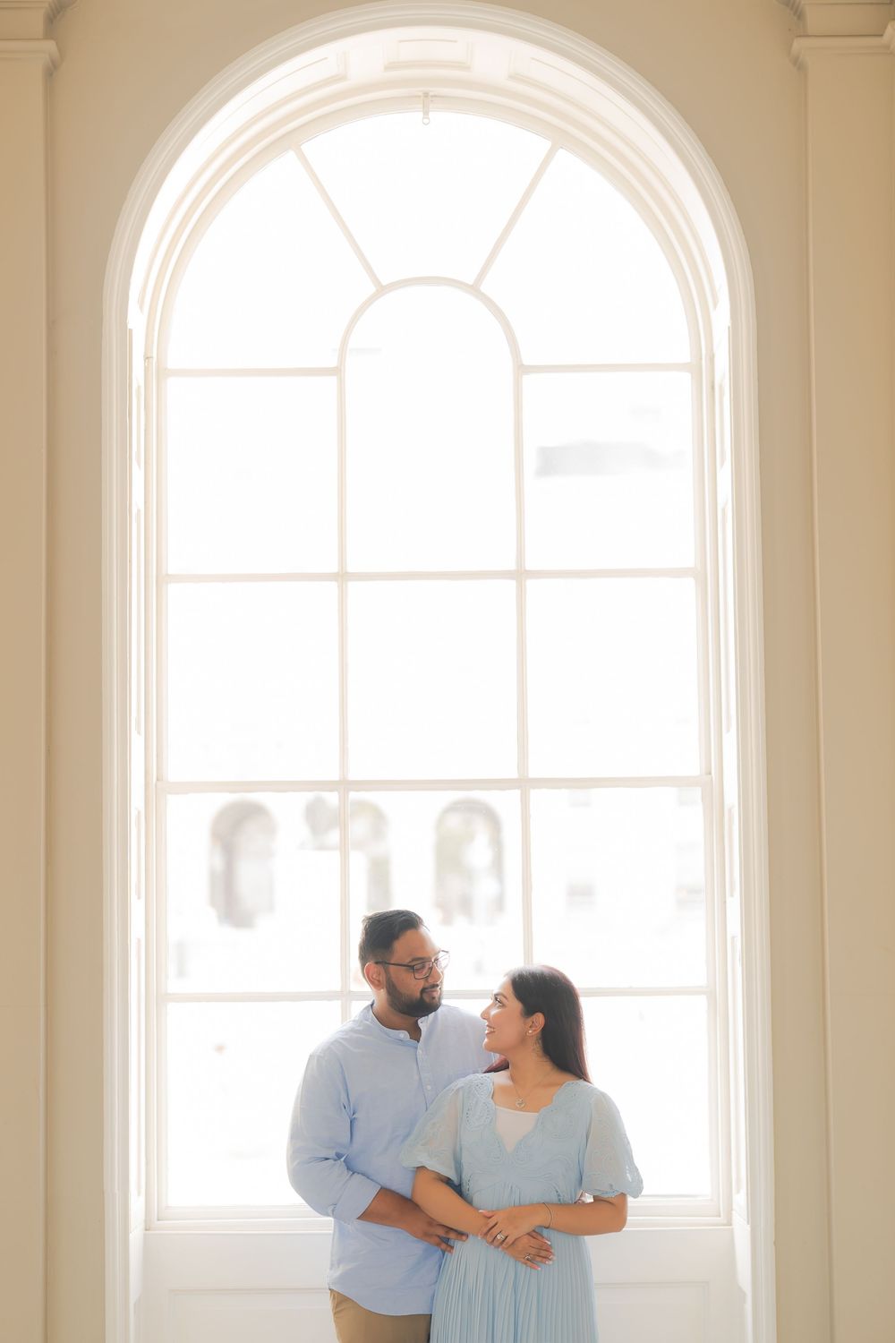 Couple in light blue outfits standing together before a large arched window in an elegant bright interior.