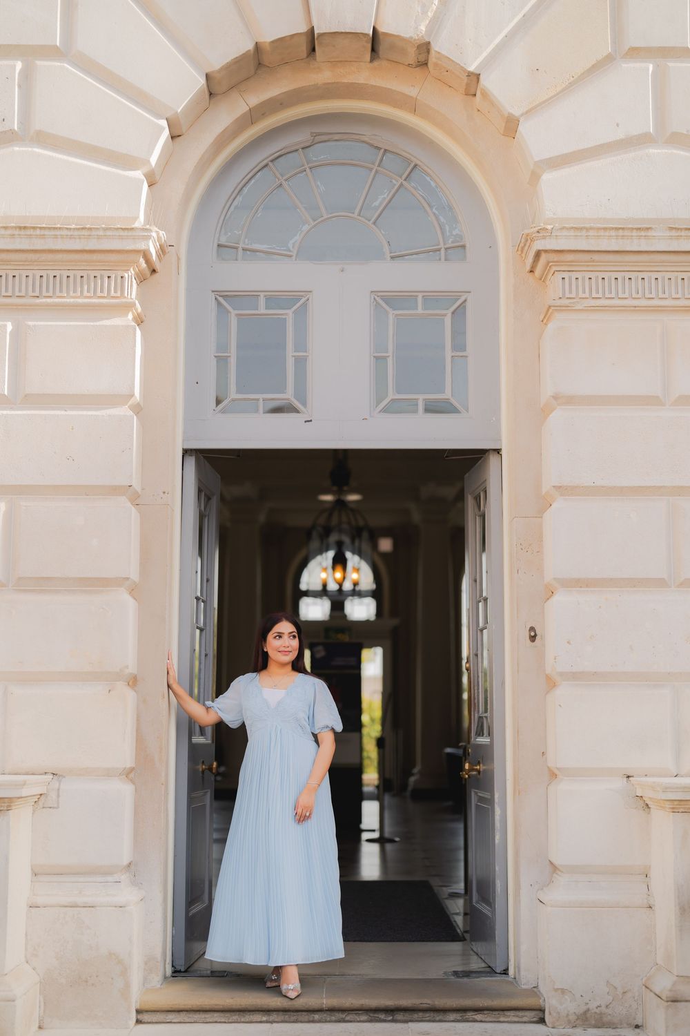 Woman in light blue maxi dress standing gracefully in an ornate arched doorway of a classic building.