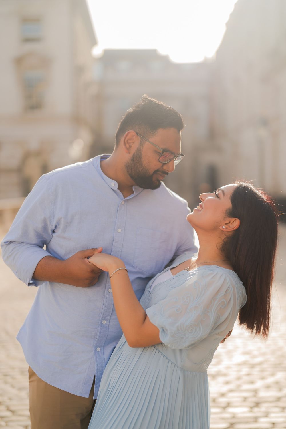 A couple in light blue outfits share a romantic moment on a sunlit cobblestone street, gazing lovingly at each other.