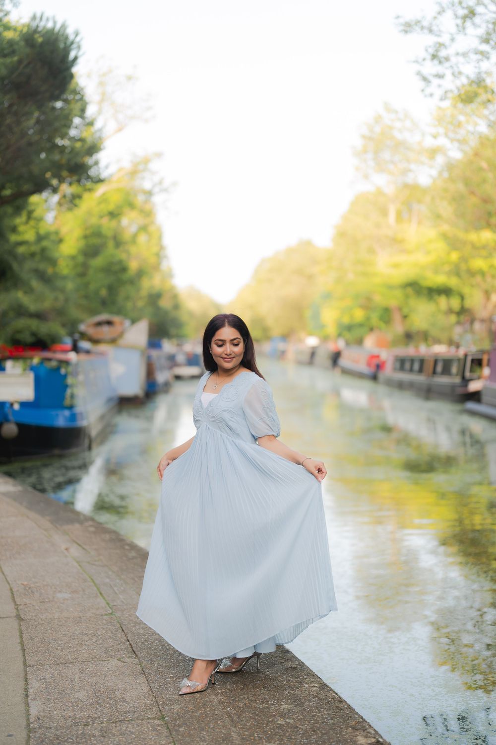 Woman in light blue maxi dress posing by a canal with narrowboats and green trees in background.