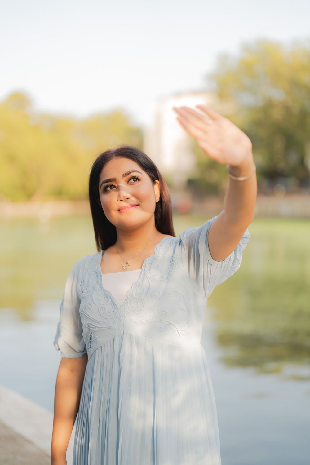 A young woman in a light blue dress waves while standing near a serene pond in a sunlit park.