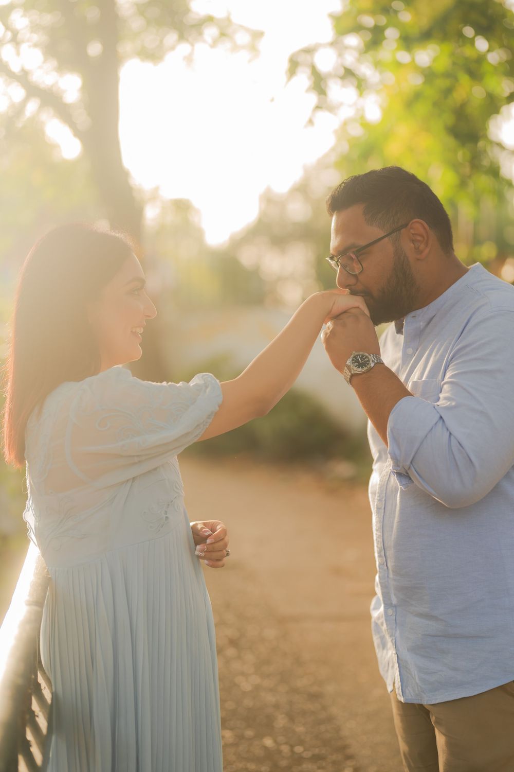 A man gently kisses a woman's hand in a sunlit park, both smiling in a romantic outdoor setting.