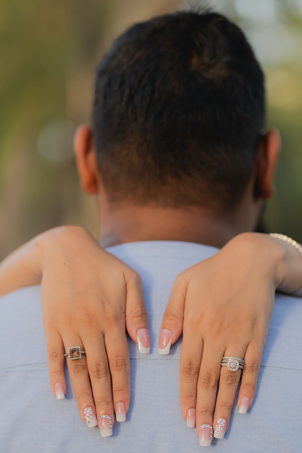Close-up of a woman's hands with French manicure and rings resting on a man's shoulders during an engagement photo.