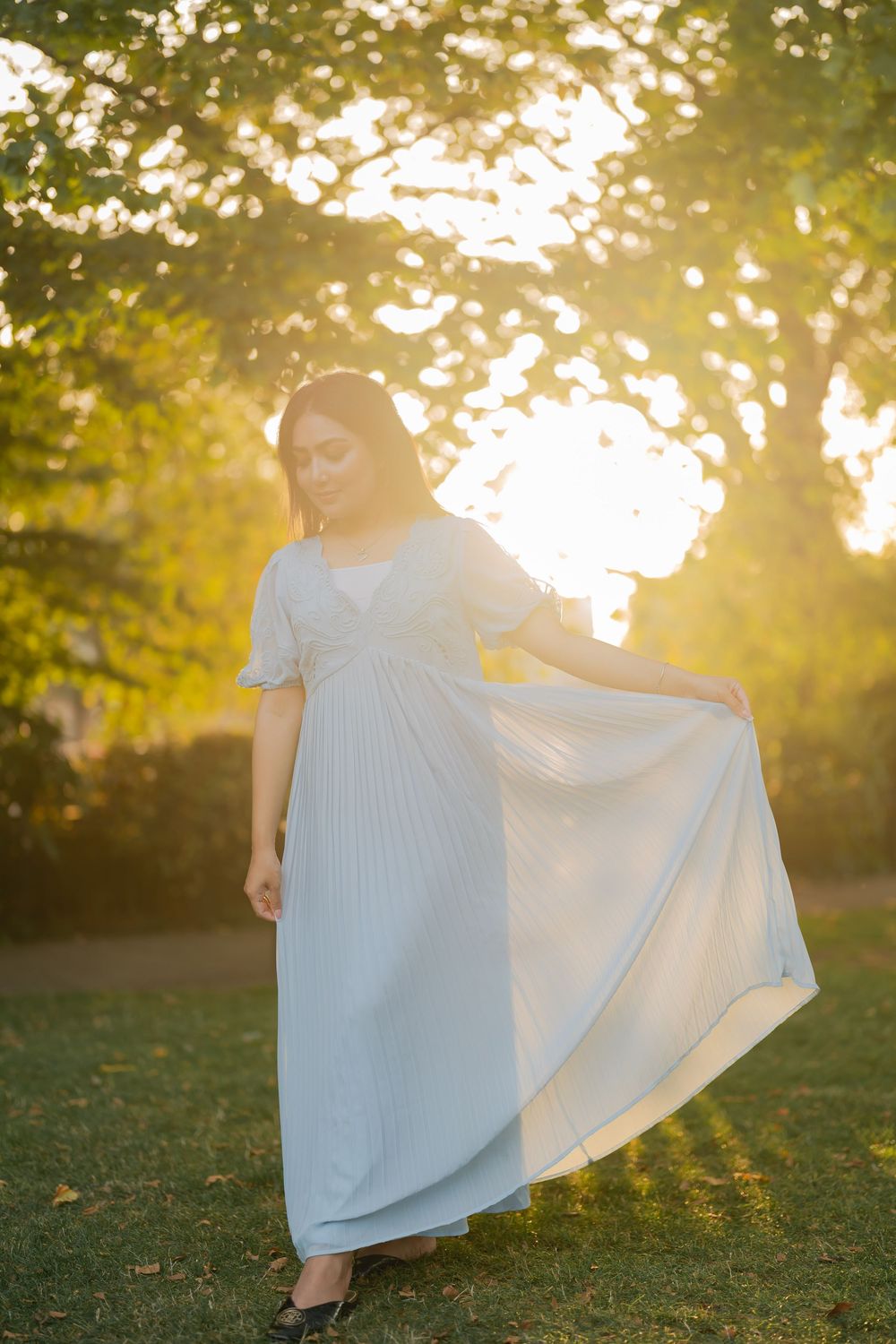 Woman in flowing light blue maxi dress posing gracefully outdoors during golden hour sunset in a lush green park.