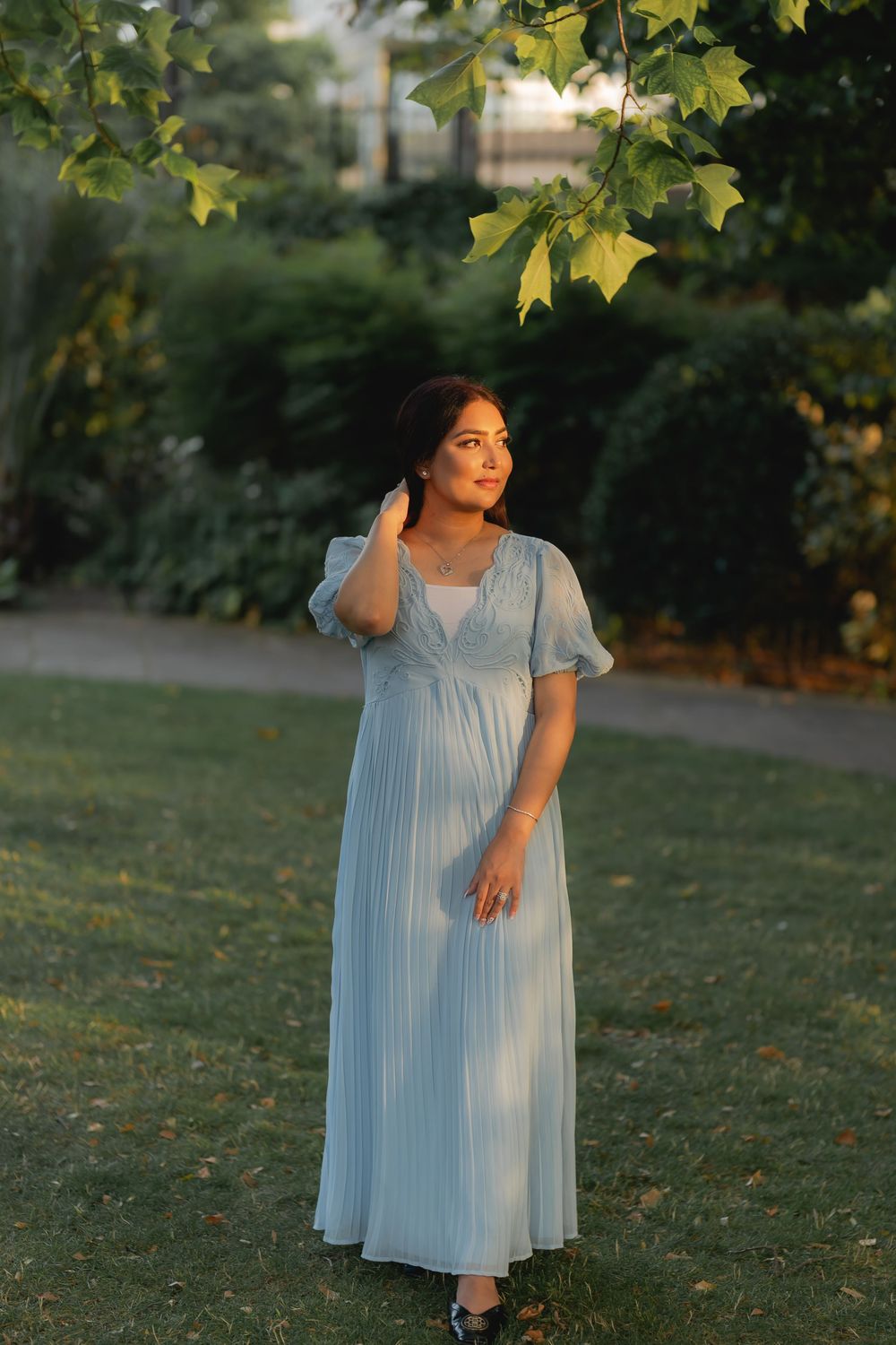 Woman in light blue pleated maxi dress standing in a sunlit garden, gazing upward near maple tree branches.