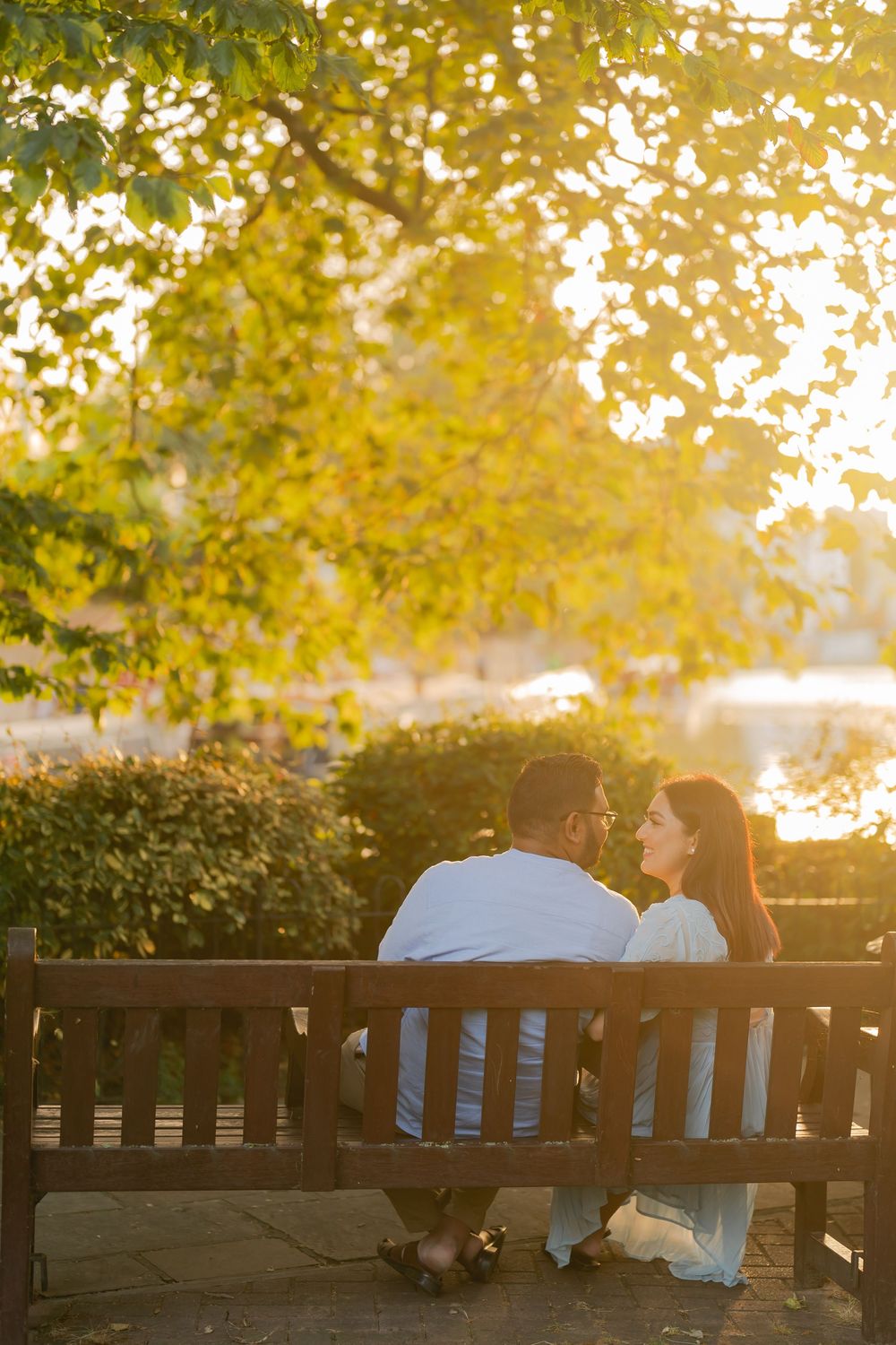 Couple sitting together on a wooden park bench at golden hour, surrounded by lush green trees near a tranquil lake.