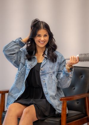 Woman in oversized denim jacket and black dress sits smiling, holding a round brush in a modern salon setting.