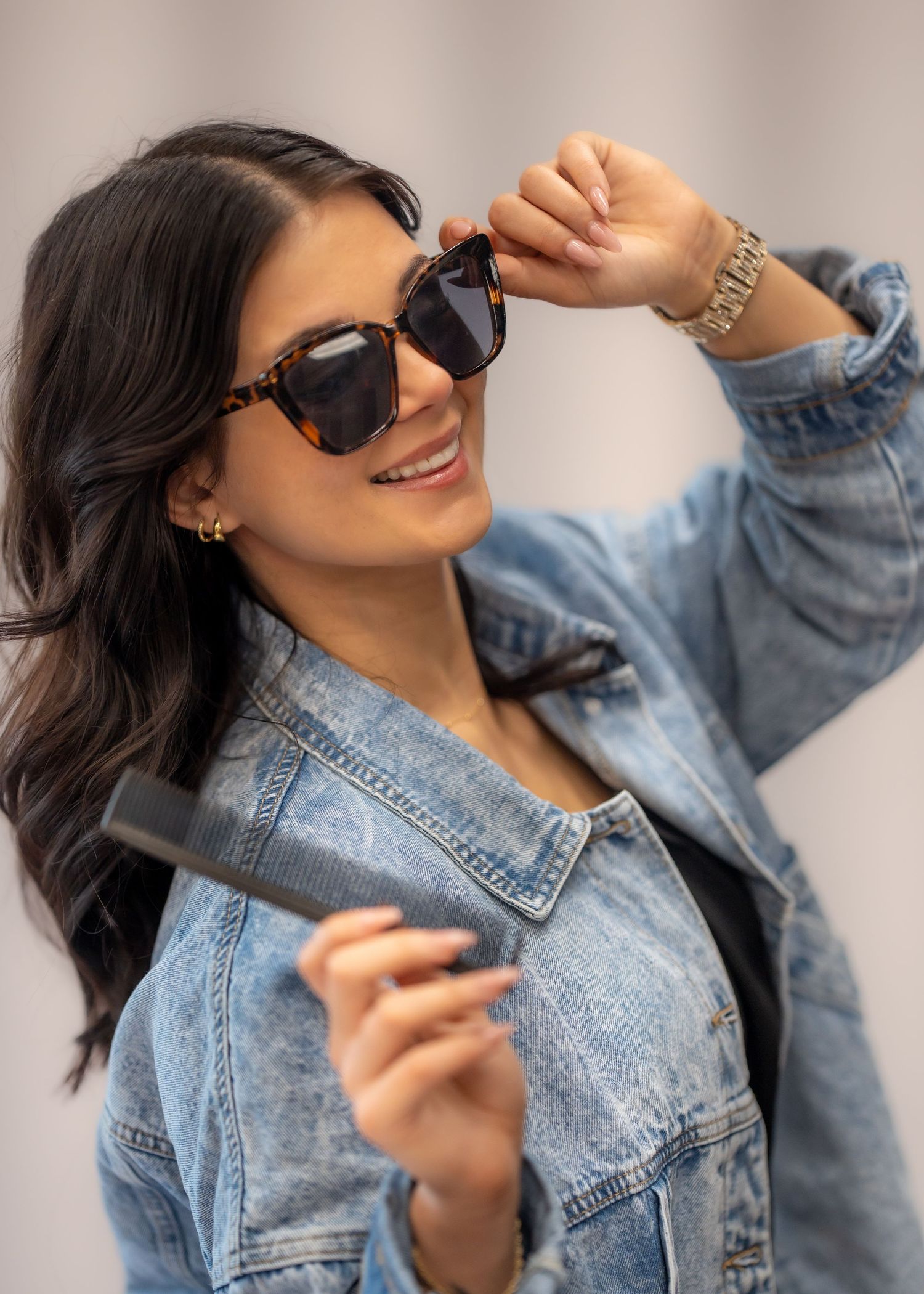 Woman in denim jacket smiling while holding tortoise shell sunglasses and a hair straightener.