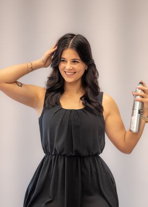 Smiling woman in black dress holding hairspray with hand in dark wavy hair, posing in a salon setting.