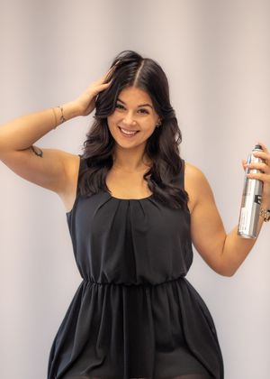 Smiling woman in black dress holding hair spray can, posing with hand in dark wavy hair in a salon setting.