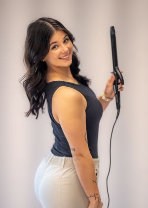 Woman smiling and holding a flat iron hair straightener, wearing a navy top and white jeans with tattoos visible.
