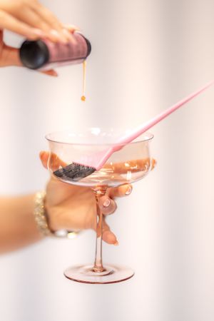 Hand holding a pink cocktail glass with a brush while pouring liquid from a bottle at a hair salon.