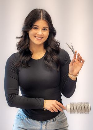 Smiling female hairstylist in black top holding scissors and a round brush in a salon studio.