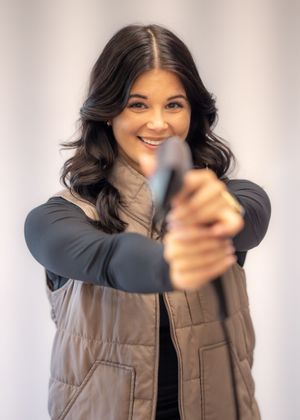 Smiling woman in tan vest aiming a handgun directly at camera in a bright indoor setting.