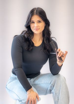 Woman in black long-sleeve top and jeans sits holding a hair comb, posing confidently against a light background.