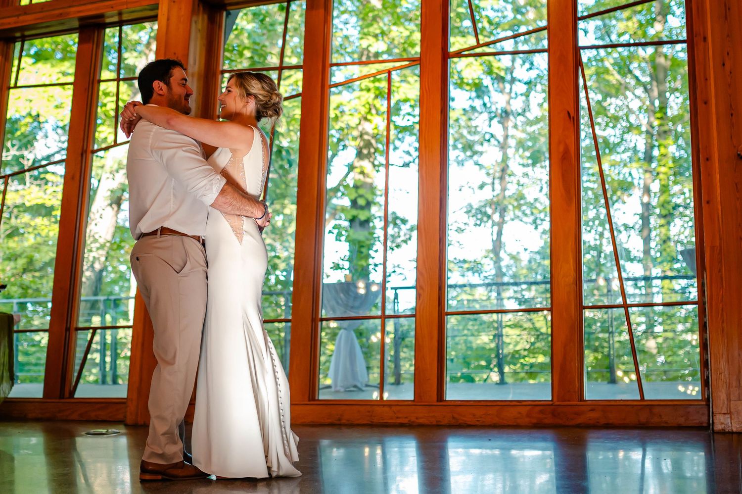 Bride and groom share romantic embrace in front of large wooden-framed windows with lush green trees in background.