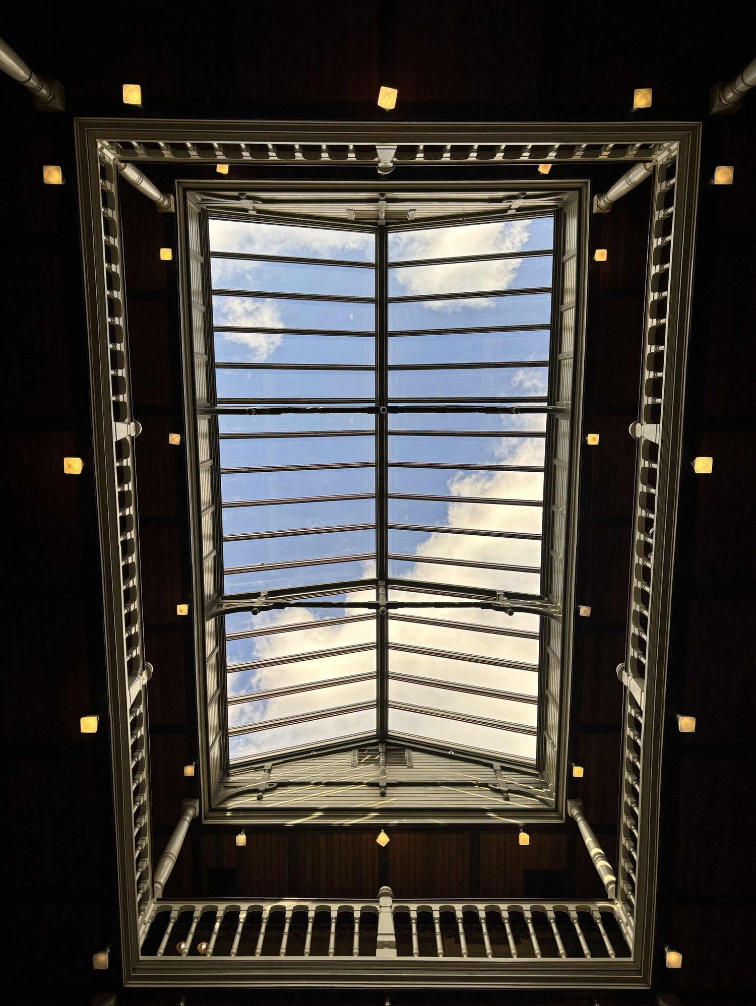Looking up through a rectangular skylight of the Beaumont Hotel with metal grid frame, revealing blue sky and clouds, surrounded by lights.