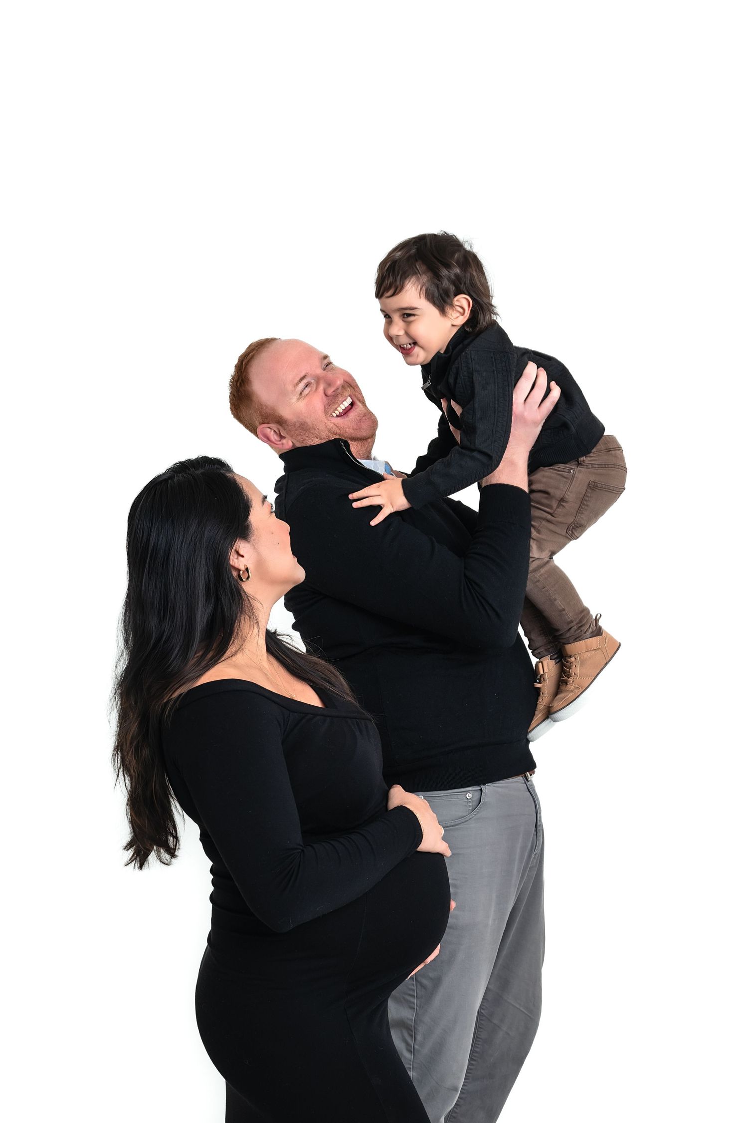 Pregnant woman in black dress stands with smiling red-haired man lifting happy young boy against white background.