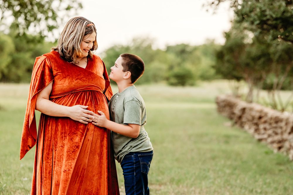 Pregnant woman in orange velvet dress with young boy touching her baby bump outdoors in green field in Fort Worth, TX.