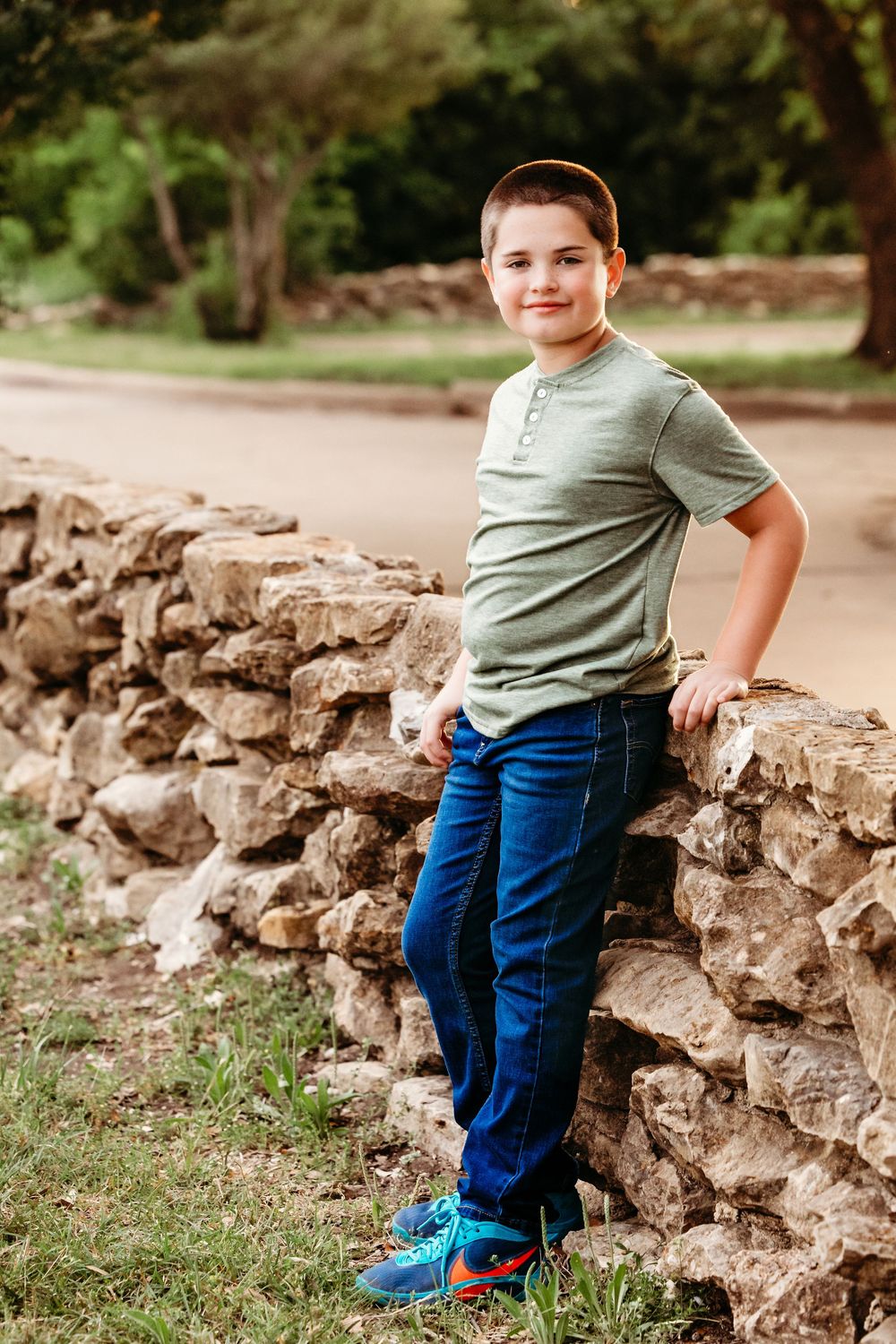 A smiling boy in a green shirt and jeans leans against a stone wall in a park setting in Fort Worth, TX.