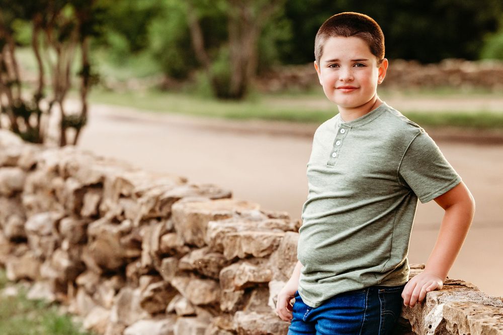 A smiling young boy in a green henley shirt leans against a stone wall outdoors in a professional portrait photo in Fort Worth, TX.