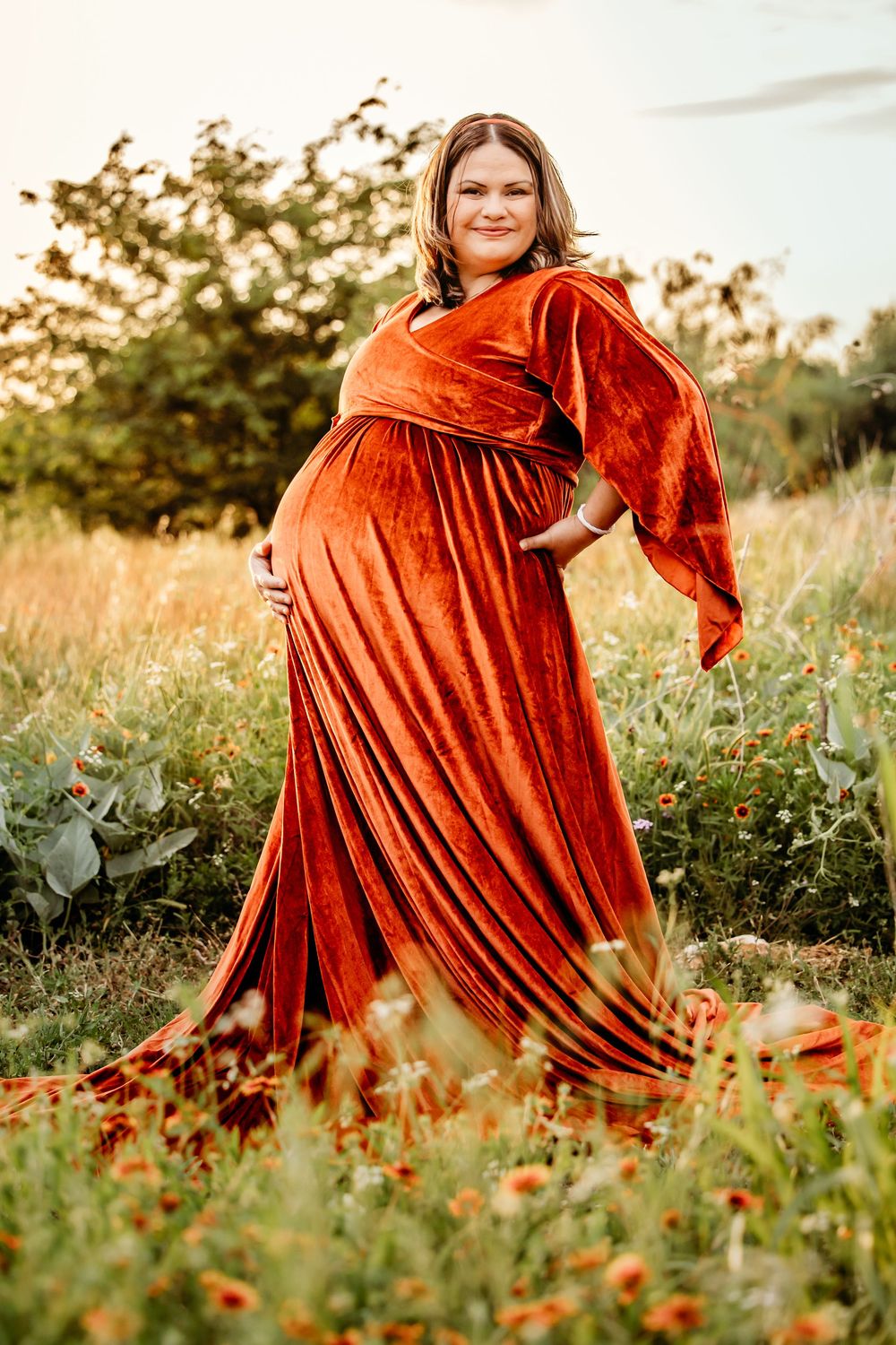 Pregnant woman in rust velvet gown standing in wildflower field at golden hour in Fort Worth, TX.