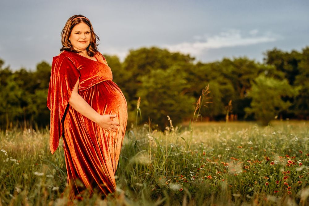 Pregnant woman in red velvet gown cradling belly in sunlit wildflower field with green trees backdrop in Fort Worth, TX.