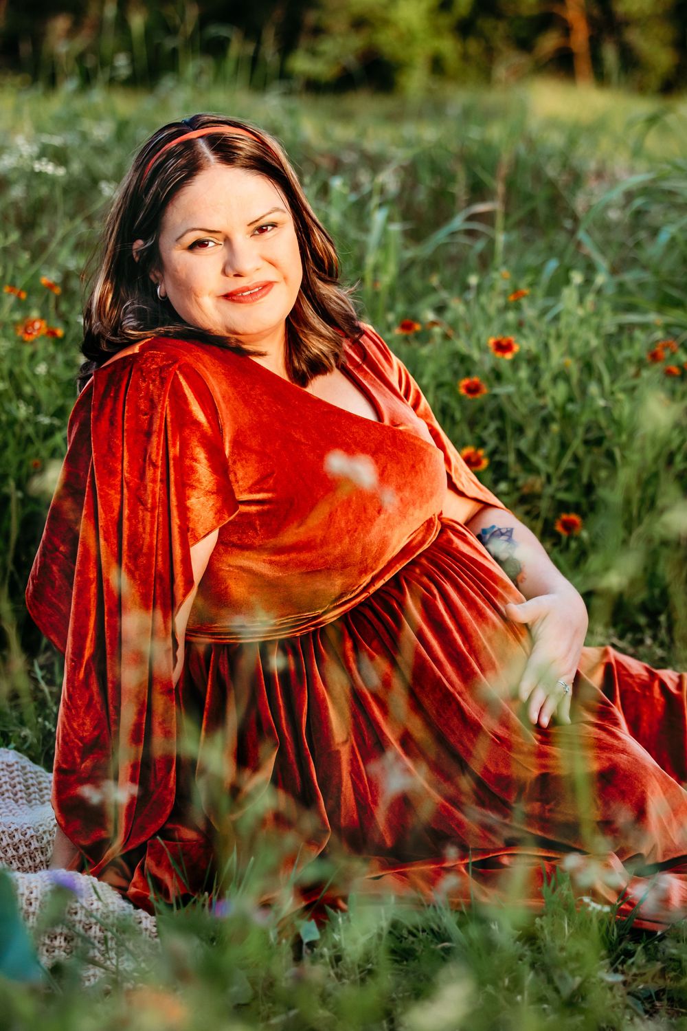 Pregnant woman in red velvet dress sitting in wildflower field, smiling warmly during outdoor maternity photo session in Fort Worth, TX.