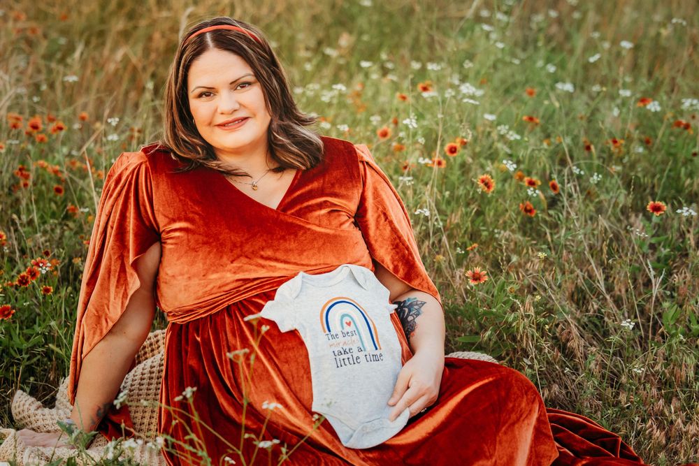 Pregnant woman in rust velvet gown sitting in wildflower field holding baby onesie during maternity shoot in Fort Worth, TX.