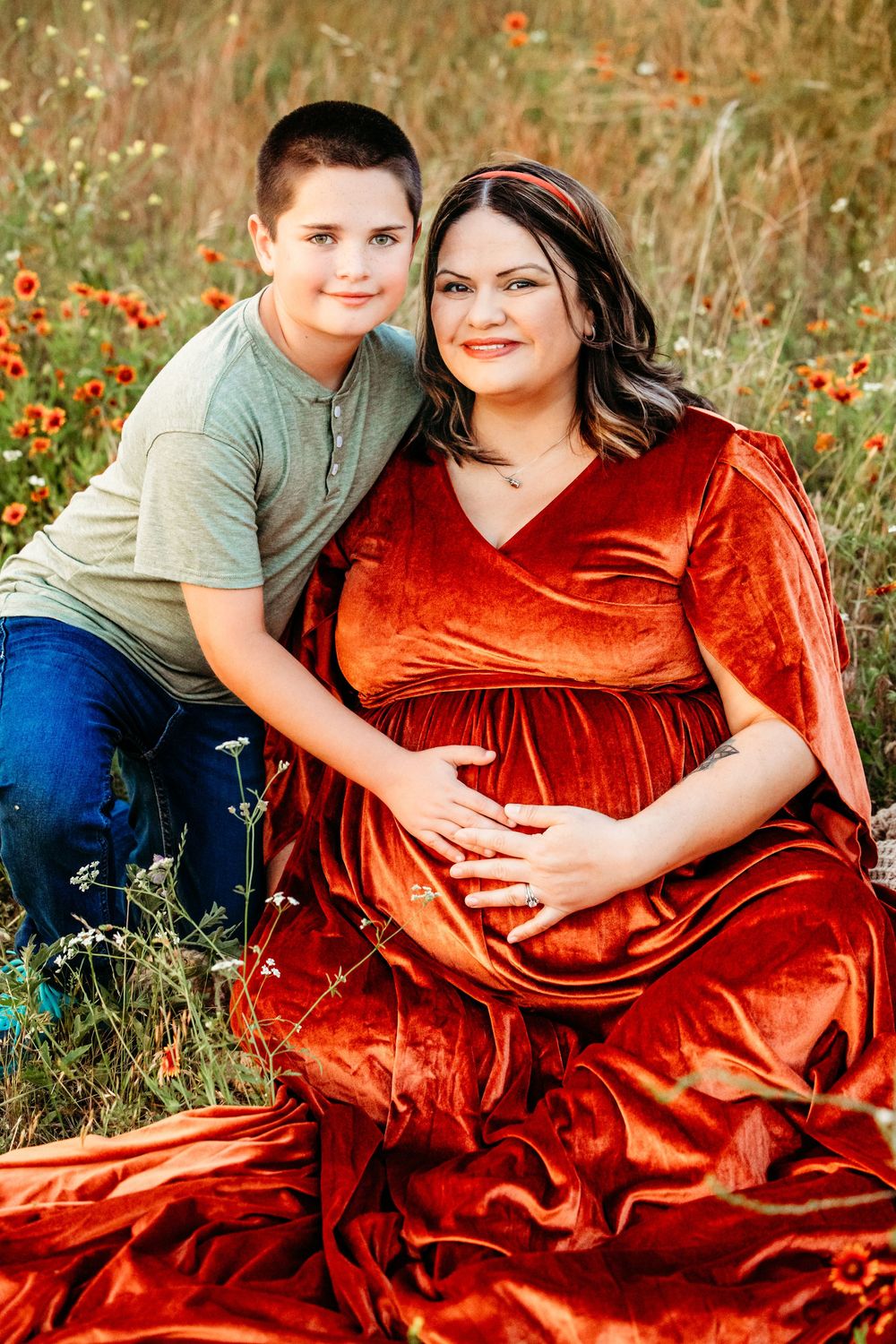 Pregnant woman in red velvet dress sits with young boy in wildflower field during maternity photo session in Fort Worth, TX.
