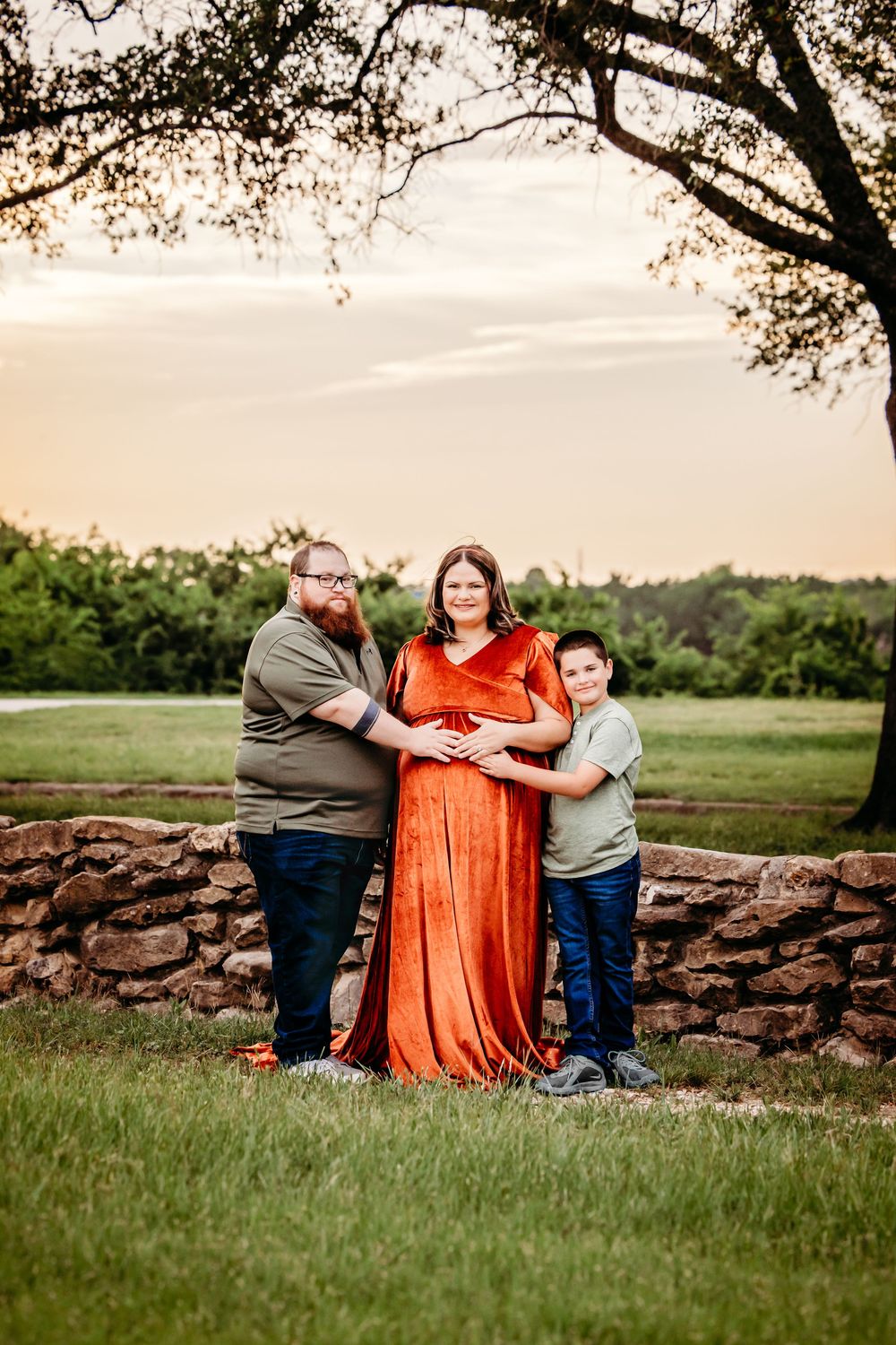 Family maternity photo with pregnant woman in rust velvet gown, partner, and child by stone wall at sunset in Fort Worth, TX.