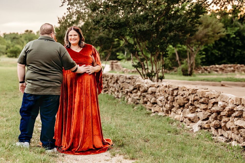 Couple sharing tender moment outdoors, pregnant woman in flowing rust velvet maternity gown near stone wall in Fort Worth, TX.