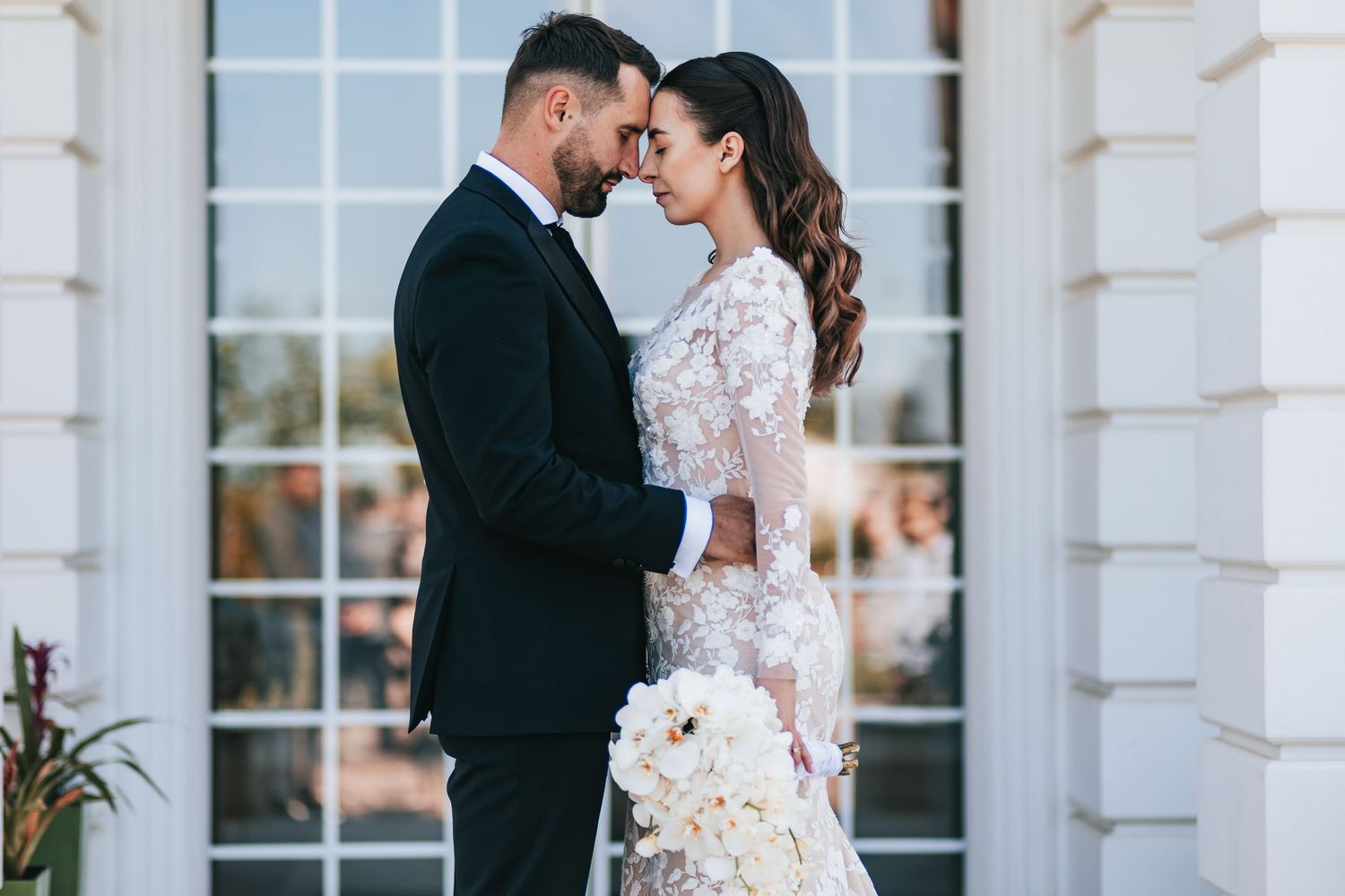 Elegant bride and groom in formal wedding attire share an intimate moment outdoors, bride holding white floral bouquet.
