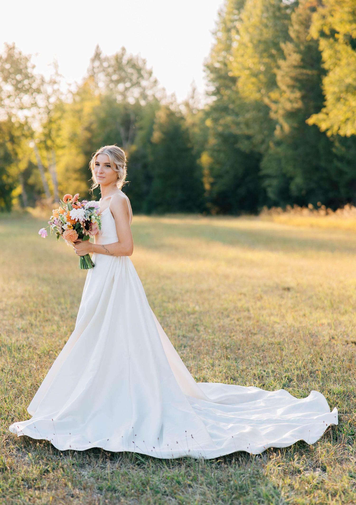 Bride in white gown with train holds colorful bouquet in sunlit meadow with lush green trees backdrop.