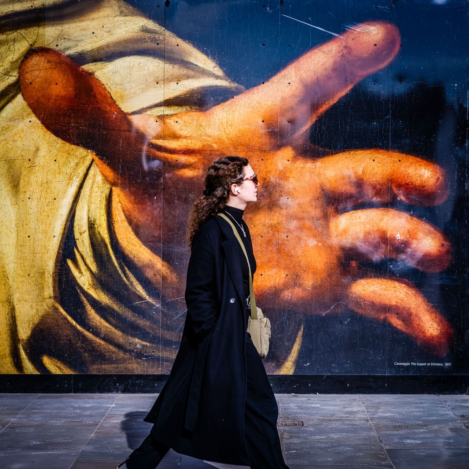 Street photography of a woman in a black coat walking past a large mural of a reaching hand