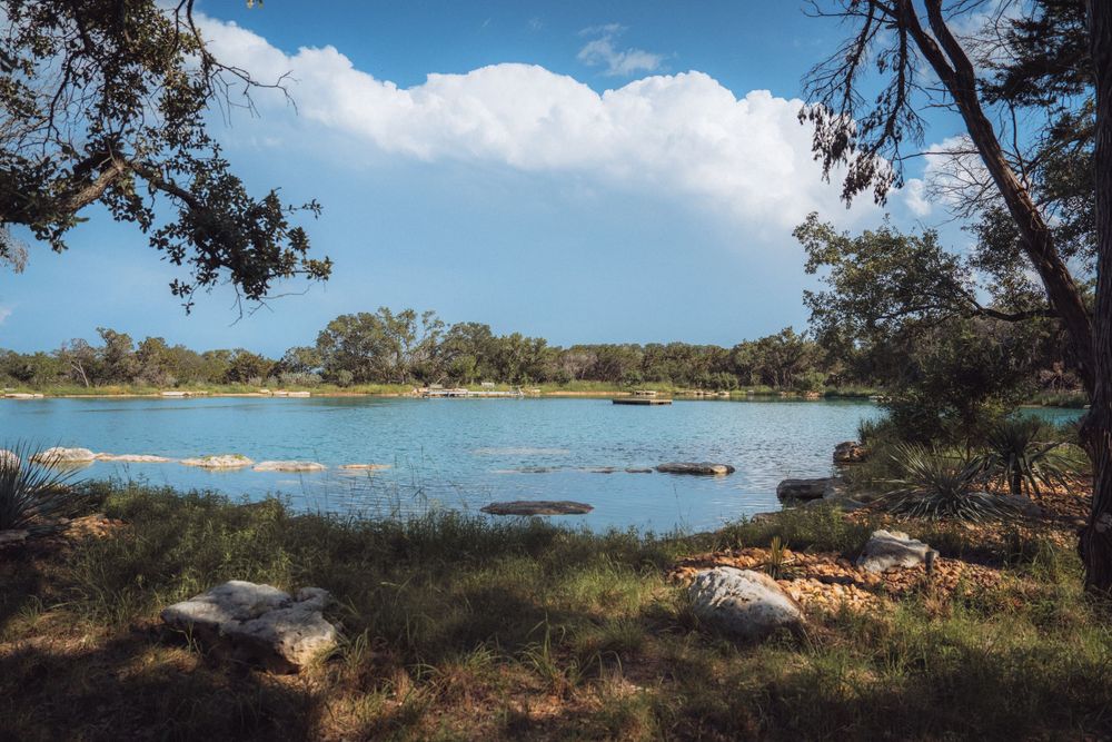 Serene lake surrounded by lush green trees and rocky shores under a bright blue sky with fluffy white clouds.