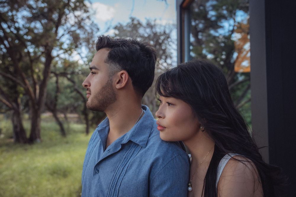 A young man and woman stand side by side outdoors, gazing thoughtfully into the distance with trees in the background.